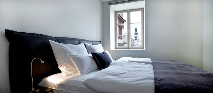 Svetla hotelska soba z belo posteljo in lesenim oknom s prečkami, skozi katero se vidi staro mestno jedro.